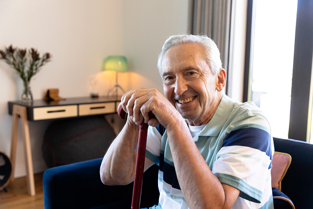 Portrait of smiling caucasian senior man with walking cane sitting on chair in living room at home. Unaltered, physical impairment, retirement lifestyle and disability concept.