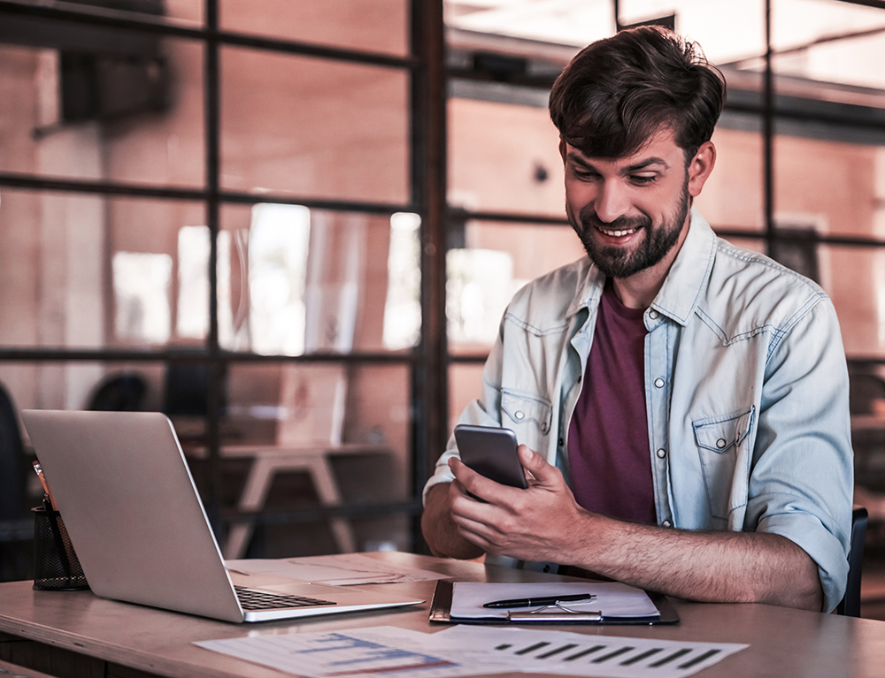 Handsome bearded businessman in casual clothes is using a smart phone and smiling while working with a laptop in office