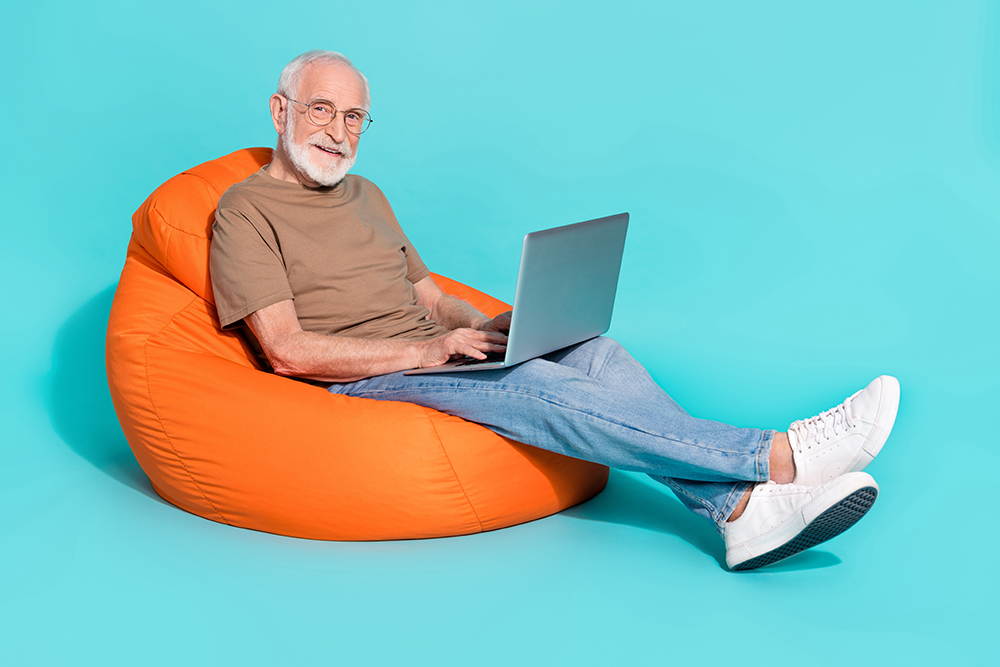 Portrait of handsome trendy cheery skilled grey-haired man sitting in bag using laptop isolated over bright blue color background