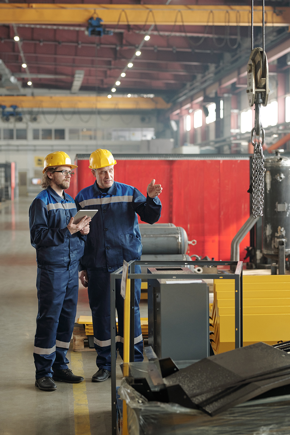 Two engineers standing in large workshop or plant and discussing new industrial equipment or machine while one of them pointing at it