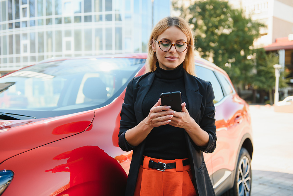 Woman using mobile phone, communication or online application, standing near car on city street or parking, outdoors. Car sharing, rental service or taxi app.