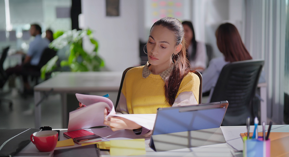 Indian gen z young adult concentrated busy business woman lady sitting chair indoor job office do day task use tablet. Focused serious modern female assistant worker do work task check paperwork