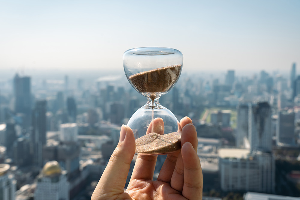 Hand holding a Hourglass with cityscape on panoramic skyline and buildings in the morning background with sun light. The concept of modern life, business, time, management and city life.