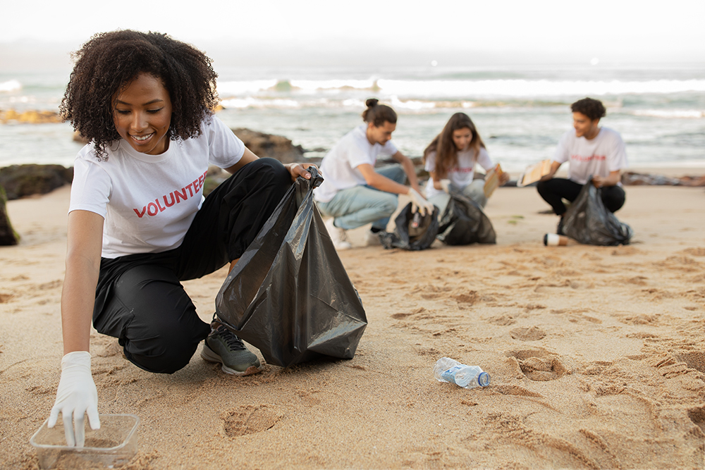 Positive young multiracial people volunteers in rubber gloves and african american lady with trash bag clean up garbage on sea beach, outdoor. Eco, environment conservation, protecting planet