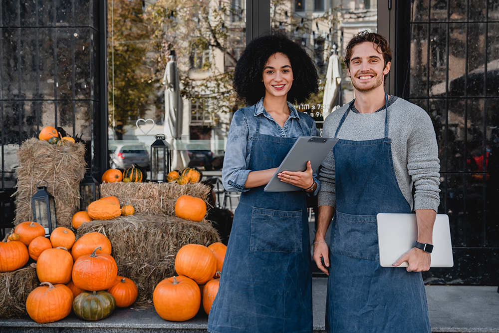 Happy young coffee shop owners waiters baristas bartenders service staff standing outdoor near their cafe with laptop and decorating pumpkins preparing for Halloween on background.