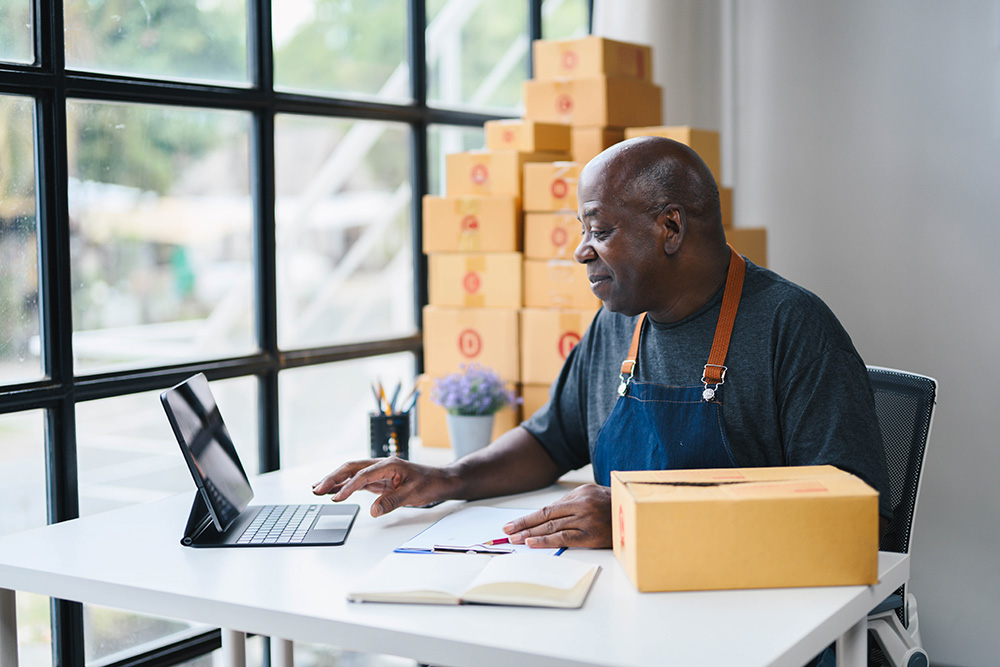 Senior man working on a tablet in a home office surrounded by cardboard boxes, representing a small business owner or e-commerce entrepreneur preparing for shipping.