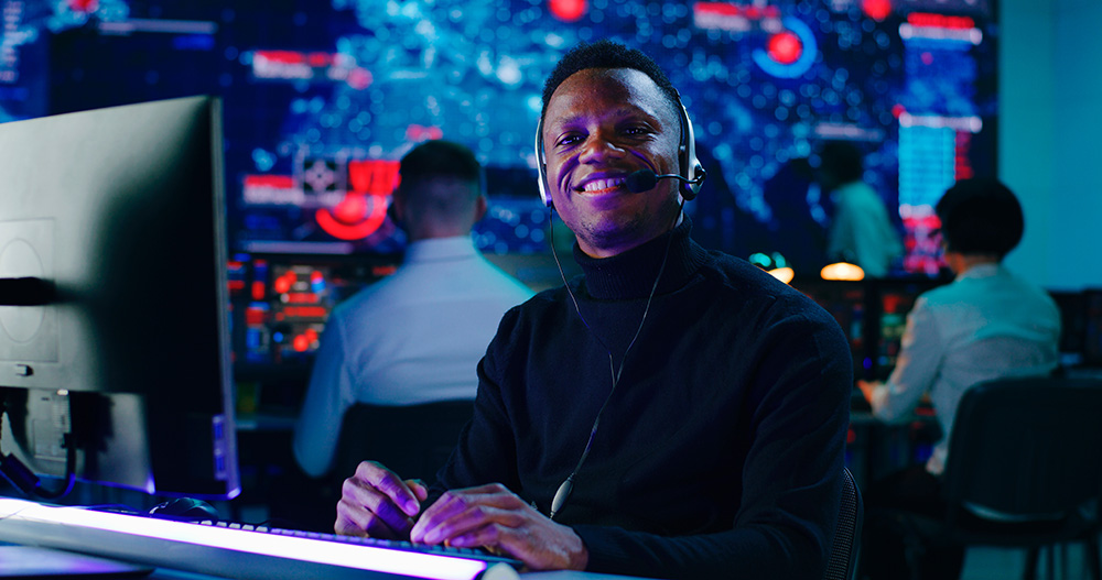 Positive black male programmer in headset using computer then smiling and looking at camera while working in office of high tech security center