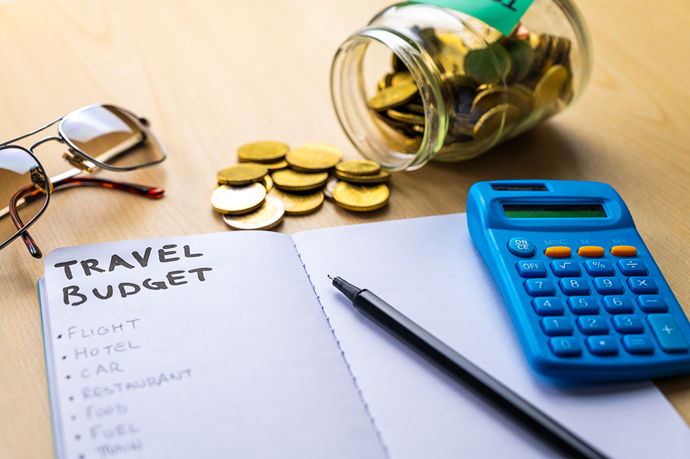 Notebook with Travel Budget and expense list, and next to it camera, glasses, money, calculator. Travel expenses.