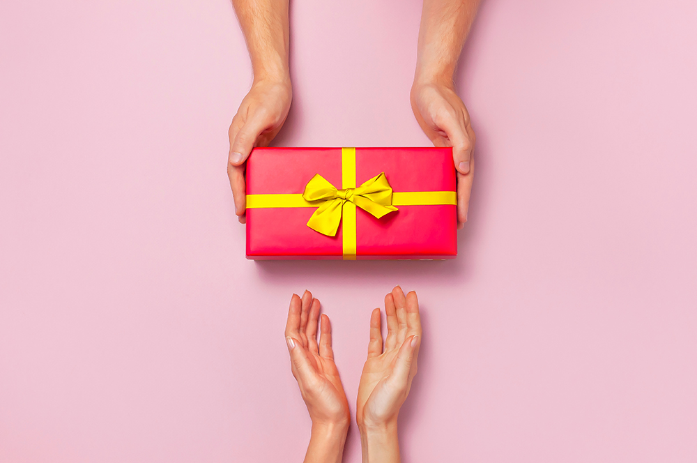 Top view of male and female hands holding red gift box with golden ribbon on pink background Flat lay. Present for birthday, valentine day, Christmas, New Year. Congratulations background copy space.