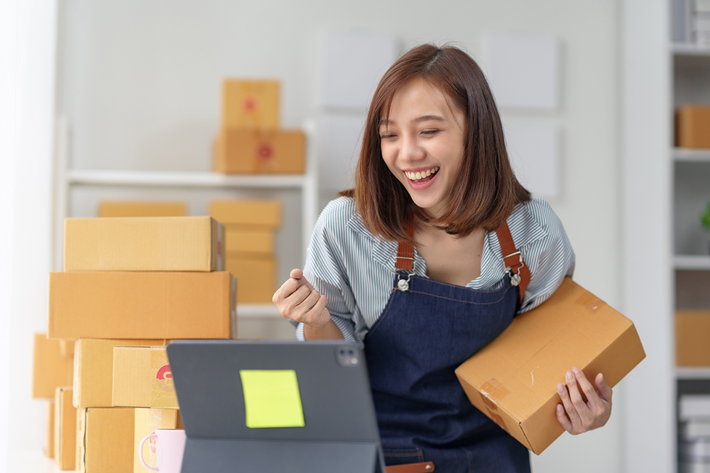 Smiling woman in apron holding box and working on laptop, surrounded by packages in a home office setting, representing successful small business.