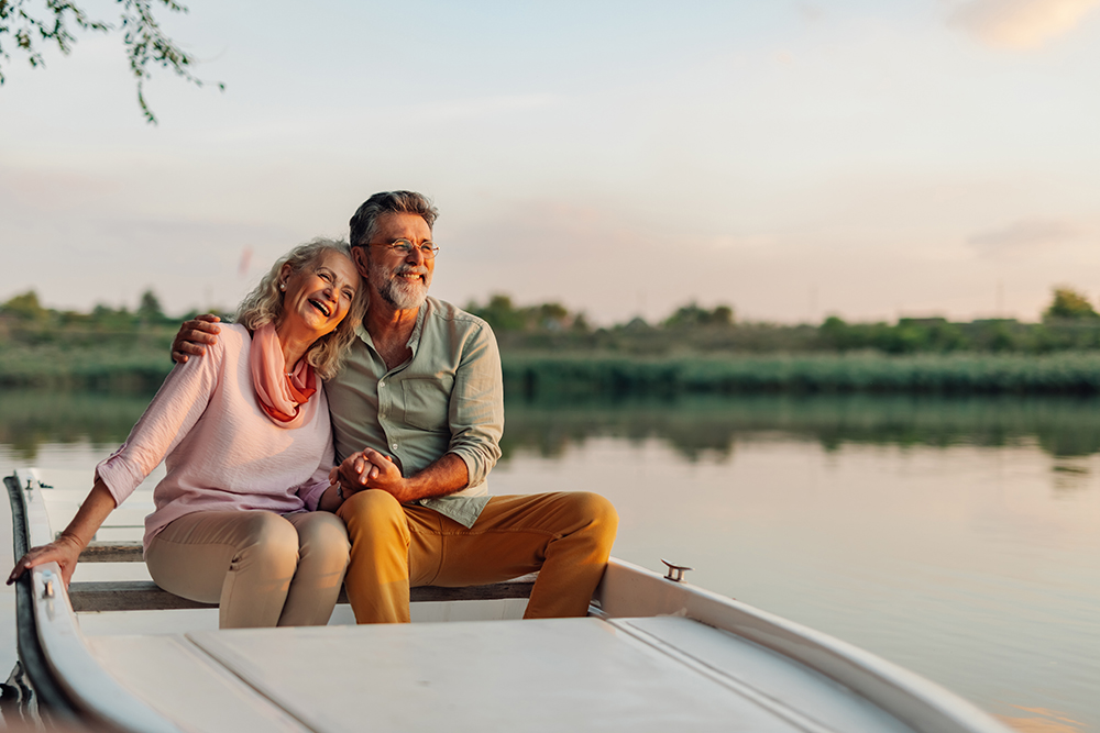 Elderly couple holding hands and laughing, enjoying a peaceful moment together while boating on a calm lake at sunset, embracing retirement and sharing a romantic experience