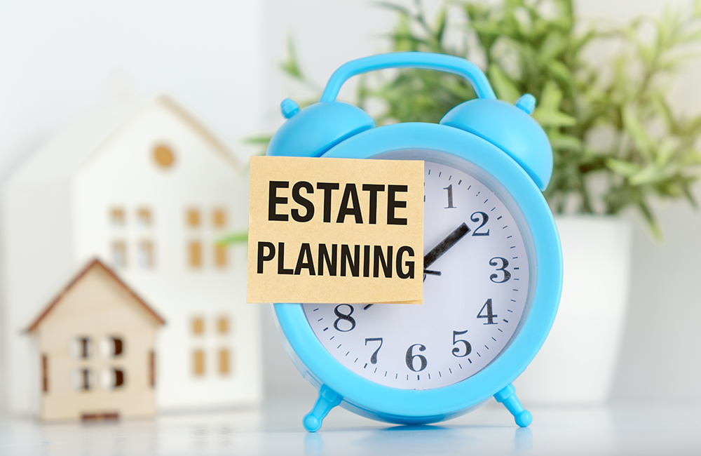 A blue alarm clock with a white paper that says Estate Planning on it. The clock is set to the time of 10:30
