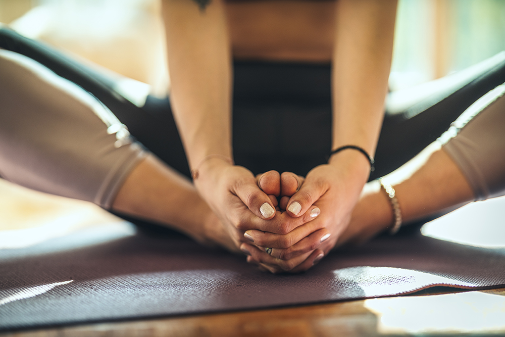 Close up of a womans hands and legs while doing yoga Lotus pose in the living room at home. She is meditating on floor mat in morning sunshine.