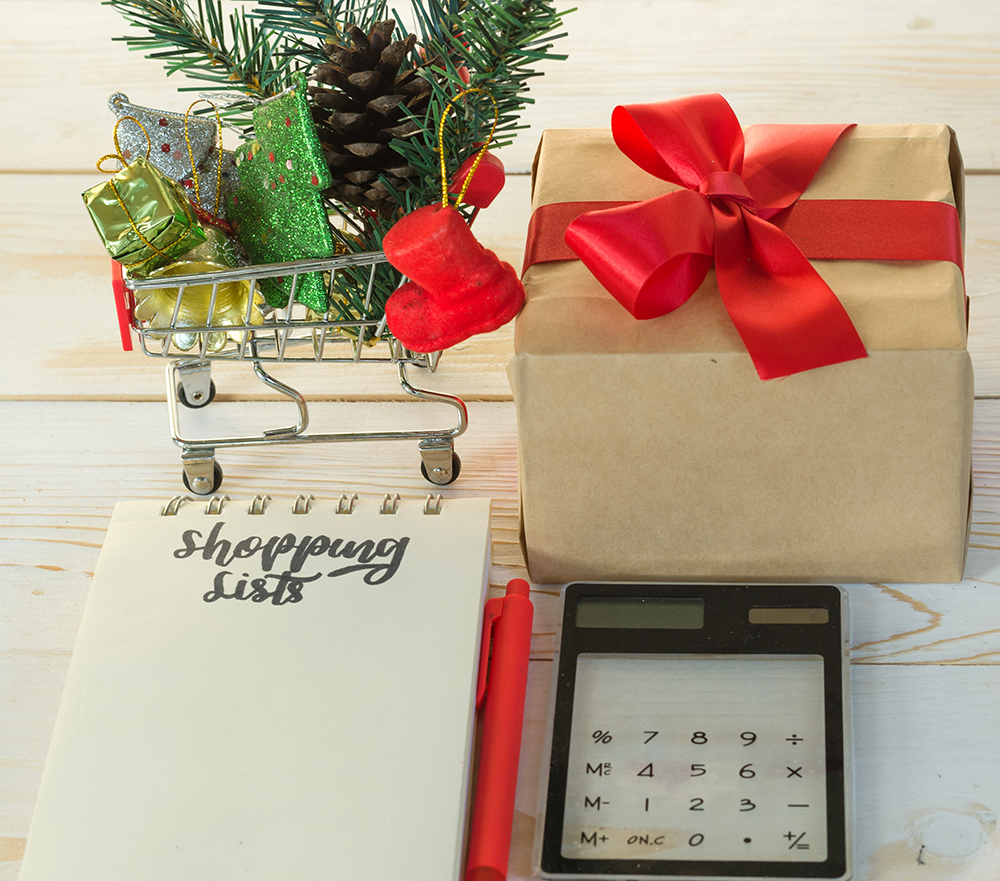 Christmas sale and shopping list for Xmas holiday sale season. Calligraphy handwriting on white notebook, supermarket cart trolley, gift box with red bow, fur, pine fruit, tree, pen and calculator.