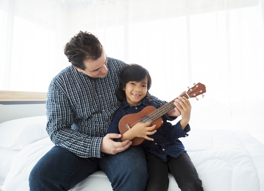 A cute handsome Asian Caucasian boy is sitting together with his dad in the bedroom while playing his ukulele