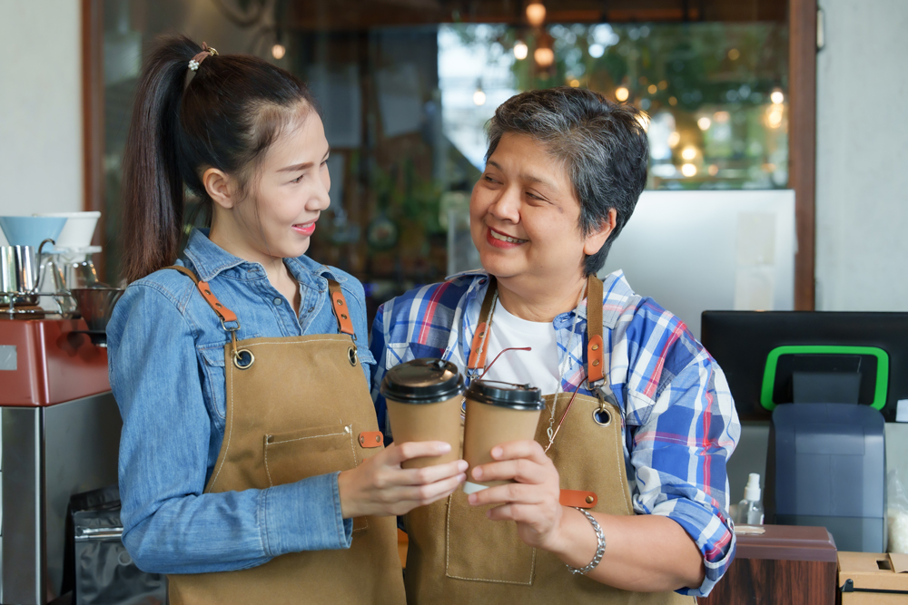 Asian elderly woman is retired. pensioner holds disposable coffee cup and clinks it with Asian daughter for promotional photo for coffee shop. The coffee shop is a family business with a happy smile.