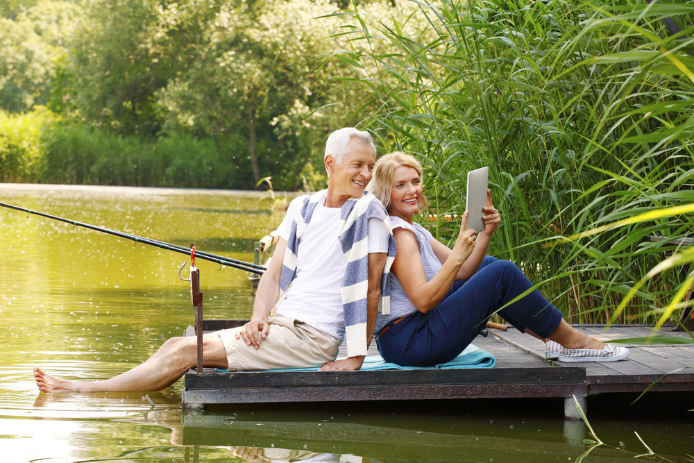 Portrait of elderly couple relaxing at nature. Senior woman and old man sitting back to back on the pier at lakeside while making self portrait with digital tablet. 