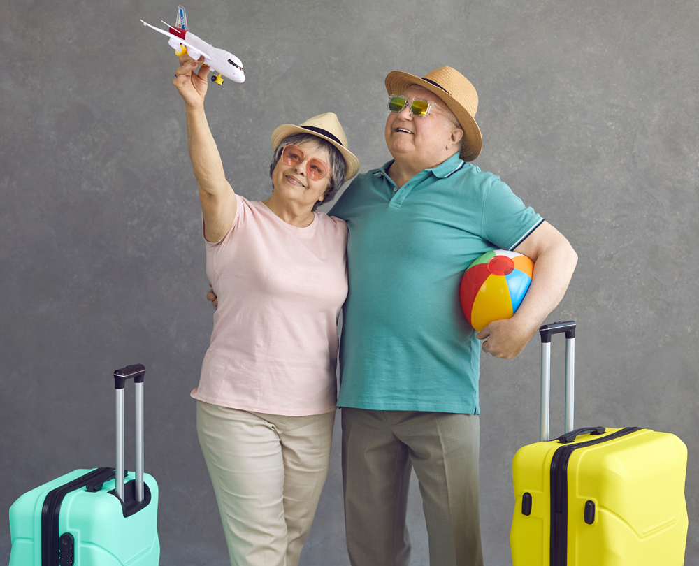 Happy old couple making dream come true. Smiling senior tourists in sunglasses and sun hats holding paper passenger airplane standing on grey studio background. Air flight and sea beach holiday