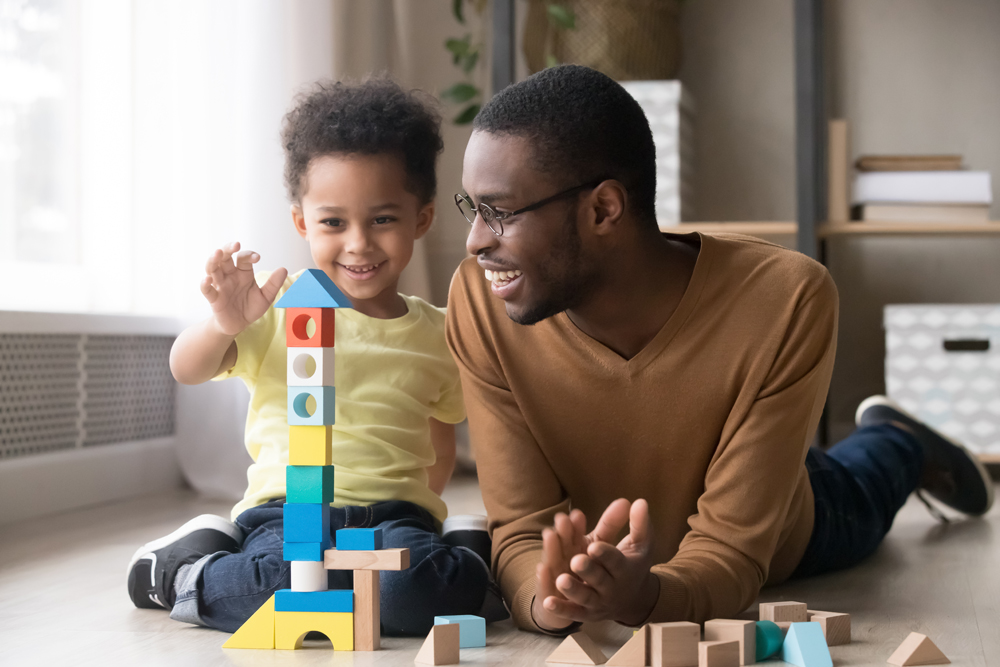 Happy cute little son playing game with black dad baby sitter building constructor tower from multicolored wooden blocks, african family father and toddler child boy having fun on warm floor at home