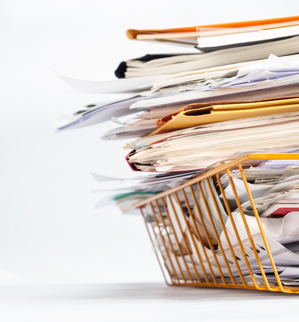 Angled shot of a yellow wire office filing or in tray, stacked high with a messy pile of documents. Copy space on white background to left and above.