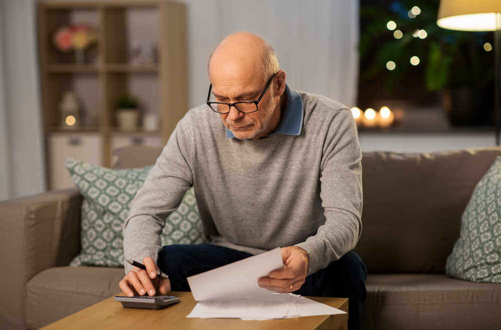 savings, annuity insurance and people concept - senior man with papers or bills counting on calculator at home in evening
