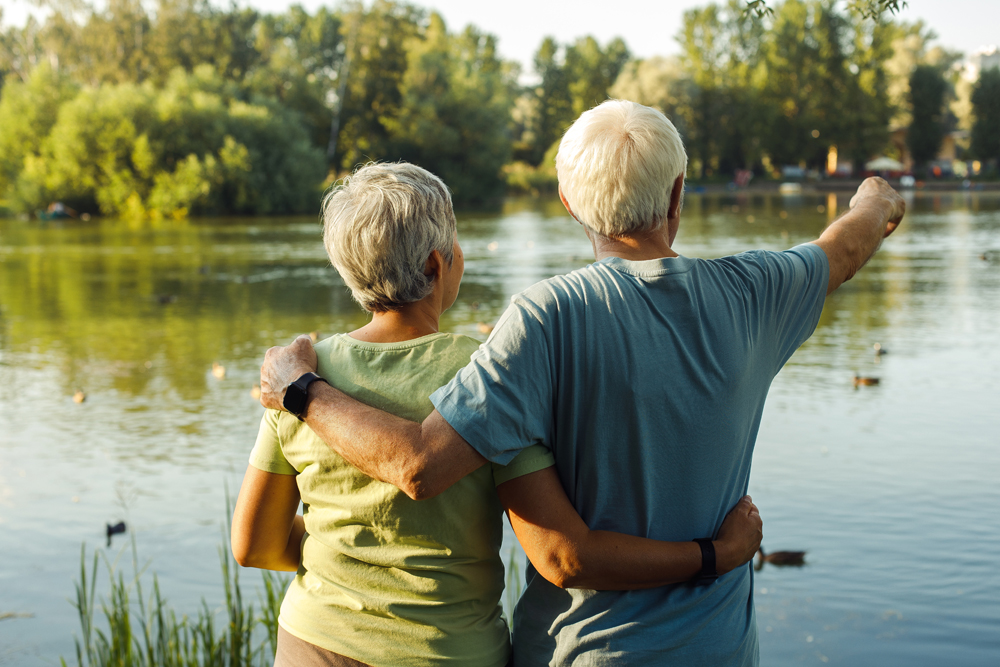 A retired couple by the lake admires the sunset on a summer day. View from the back. Golden age. Love and tenderness. Lifestyle and old people concept.