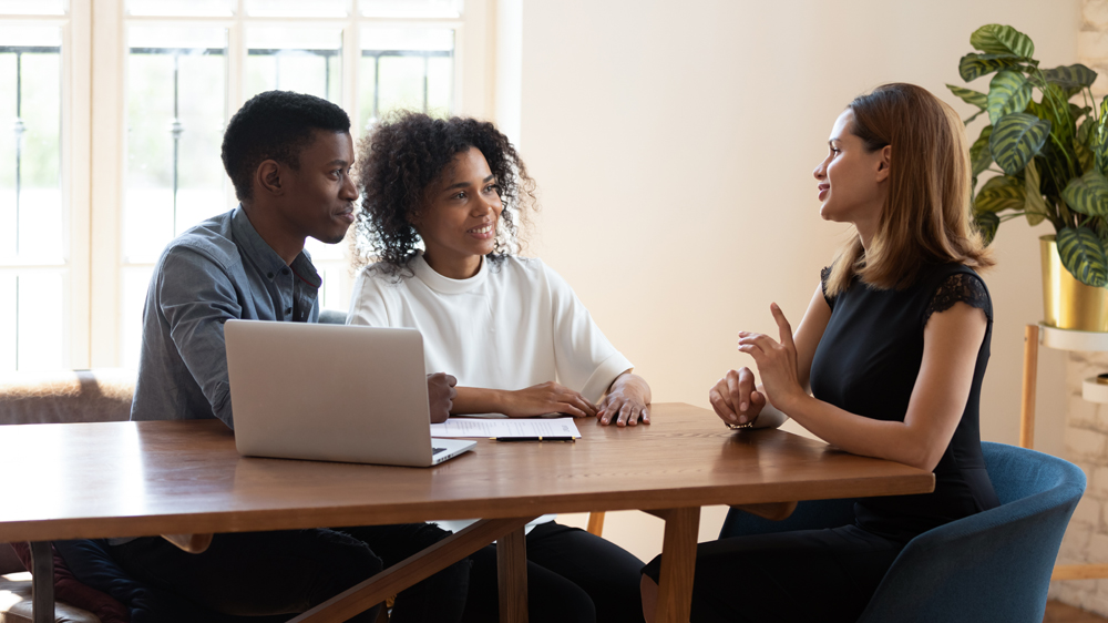 Female broker makes offer to young African ethnicity married couple during meeting in company office. Family taking loan in bank, listening advisor, real estate agent and customers negotiating concept