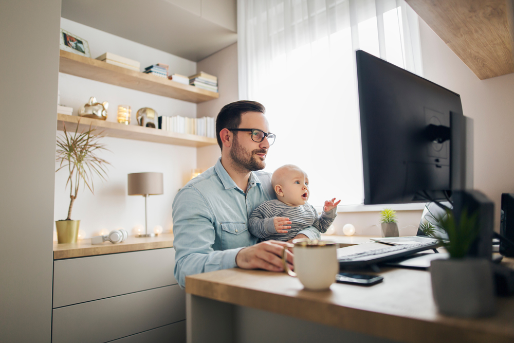 Young parent working from home while holding his baby boy on his lap while they both watch to the screen.