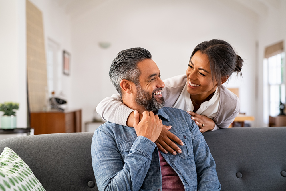 Smiling ethnic woman hugging her husband on the couch from behind in the living room. Middle eastern man having fun with his beautiful young wife on the couch. Mid adult indian man with latin woman.