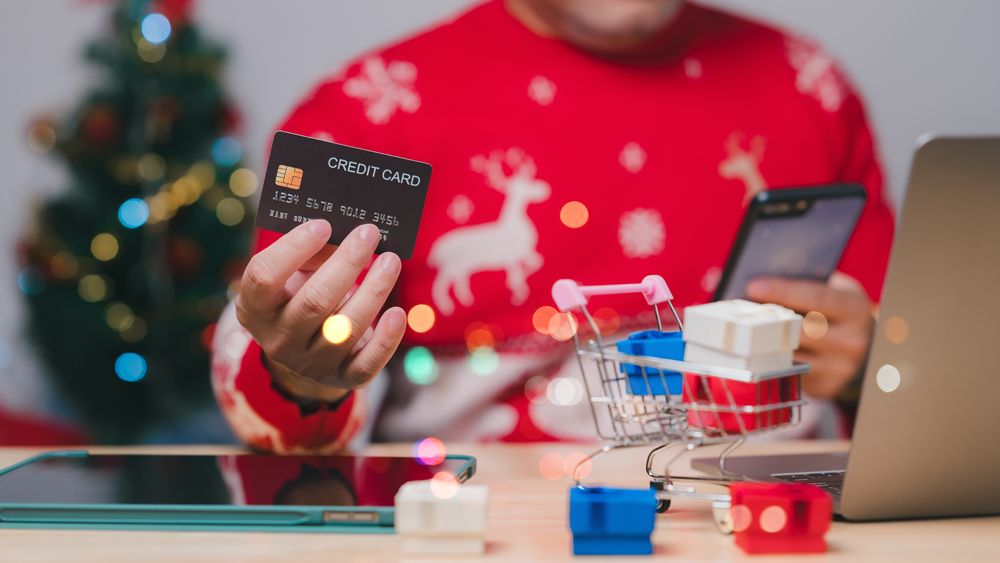 Capturing the holiday spirit, a person in a Christmas sweater holds a credit card, preparing for online shopping by phones, surrounded by festive decorations and a mini shopping cart