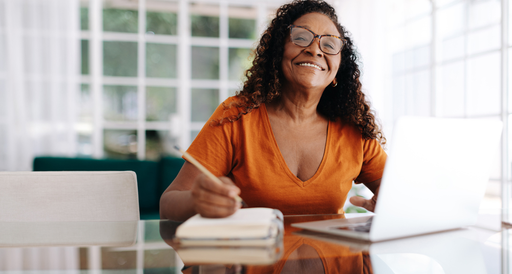 Black woman happily planning her retirement from the comfort of her own home. Mature woman sitting at a table with her laptop, writing her goals and dreams for the future in a journal.