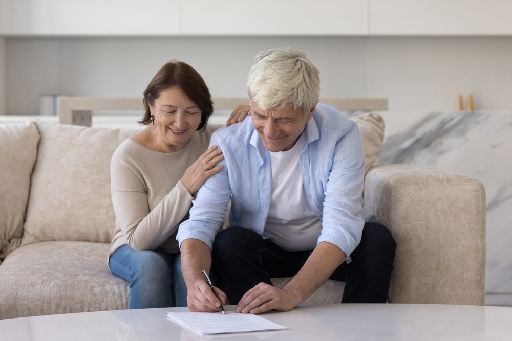 Old man writes, fill application form, buy services for comfort living, prepare formal document for lawyer, make testament, accepting terms and conditions of legal papers sit on sofa with wife at home