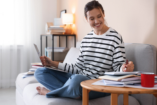 Smiling student with laptop, notebook and pen preparing for exam on sofa at home