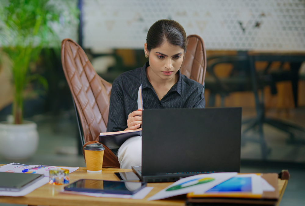 Indian young adult gen z woman sitting on sofa couch use laptop look screen do online work check bills through e-bank app. Serious female worker jotting information make notes in copybook at office
