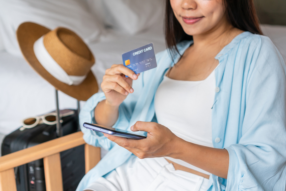 Happy traveler Asian woman holding credit card while using mobile phone with travel accessories in hotel room. Online shopping and travel, cashless society, technology and lifestyle concept