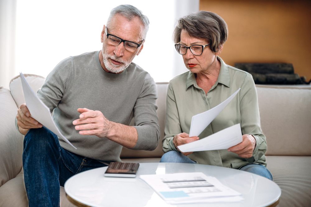 mature couple fill tax form, holding current expense document in hands, while sitting on couch at home. wife and husband receive utility bill, checking banking account score