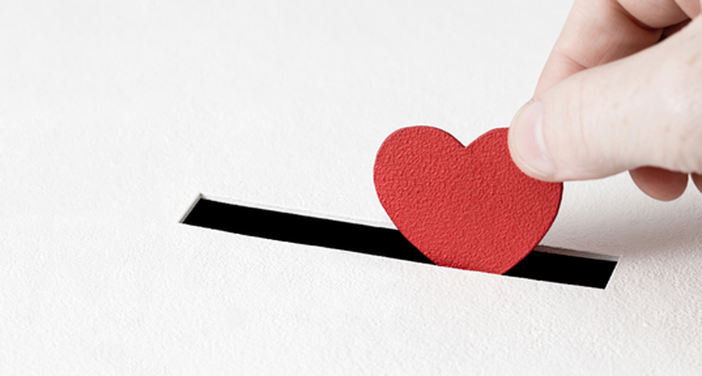 Red heart symbol is put by persons hand into slot of white donation box. Concept of donorship, life saving or charity, sincere devotion to faith. Close-up shot, horizontal banner with copy space