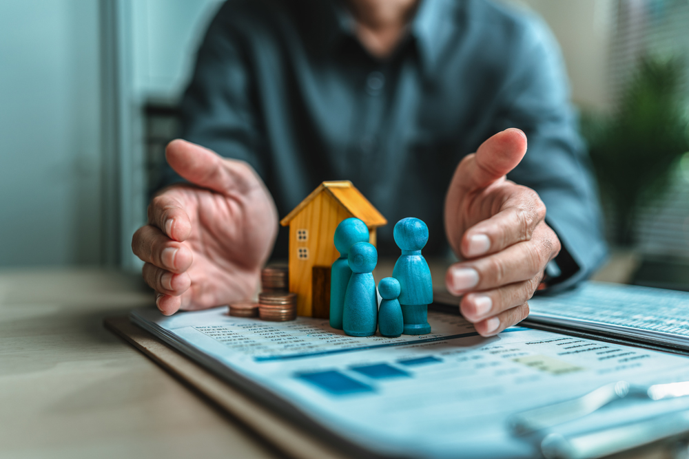 Businessmans hand gestures to protect money, house, wooden dolls represent family. Image represents the concept of asset protection, emphasizing safeguarding financial investments and property