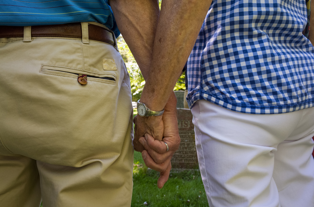 A couple of seniors holding their hands in a cemetery, mourning in front of a tombstone. 
