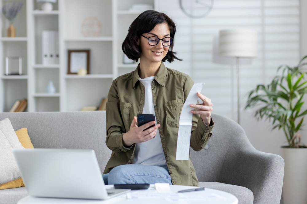 A young Asian woman with glasses, smiling while managing bills and finances using a smartphone and laptop in a well-lit, cozy living room.