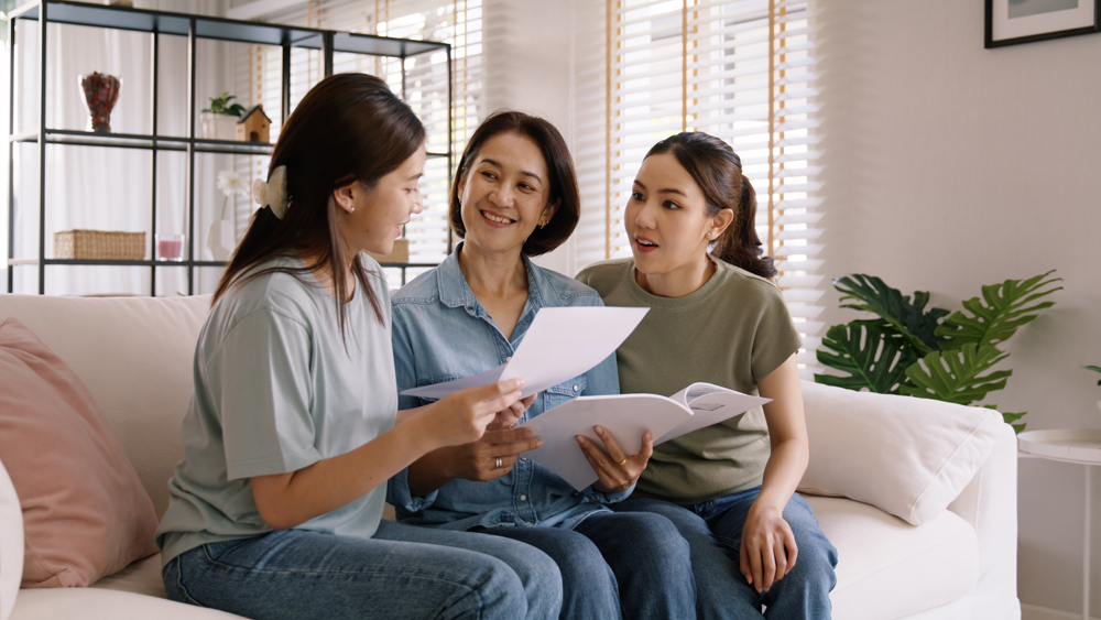 Buy mediclaim health care life insure for older mum. Young adult woman asia people grown up child give gift to middle aged mom reading protect cover saving plan paper smile laugh sitting at sofa home.