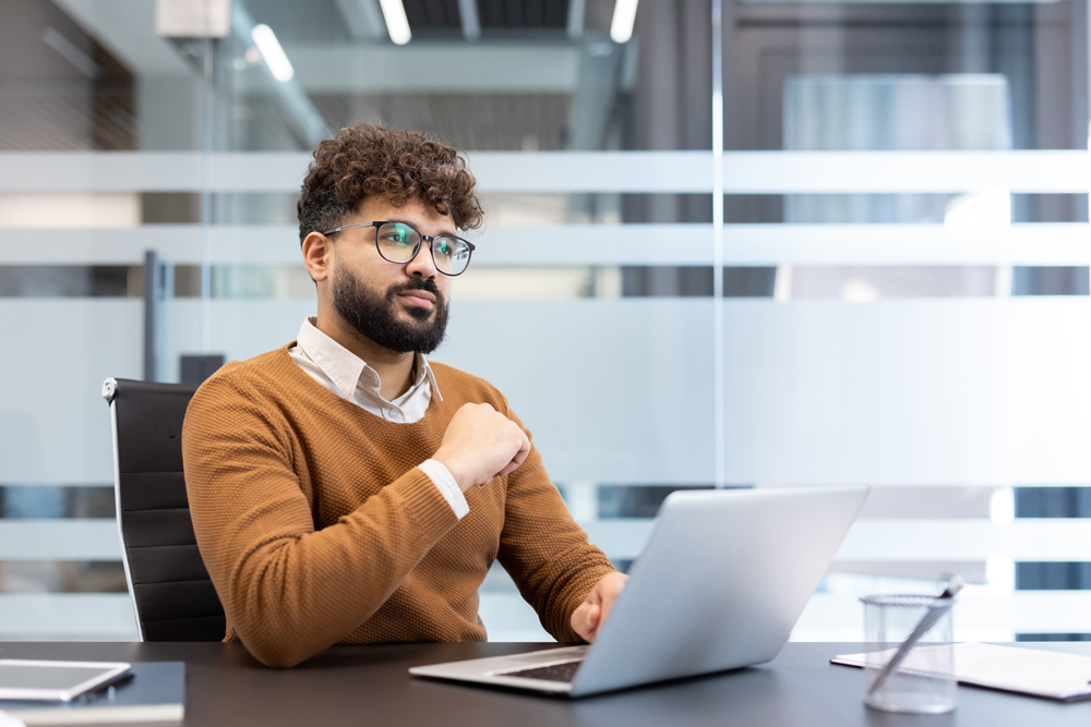 Young male professional wearing glasses and a warm sweater concentrating on a computer screen, reviewing ideas and information in an contemporary office setting