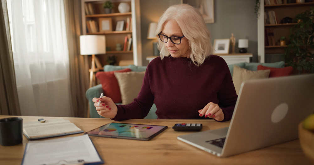 A person is sitting at a wooden desk in a home office. They are using a tablet and calculator while reviewing documents related to life insurance.