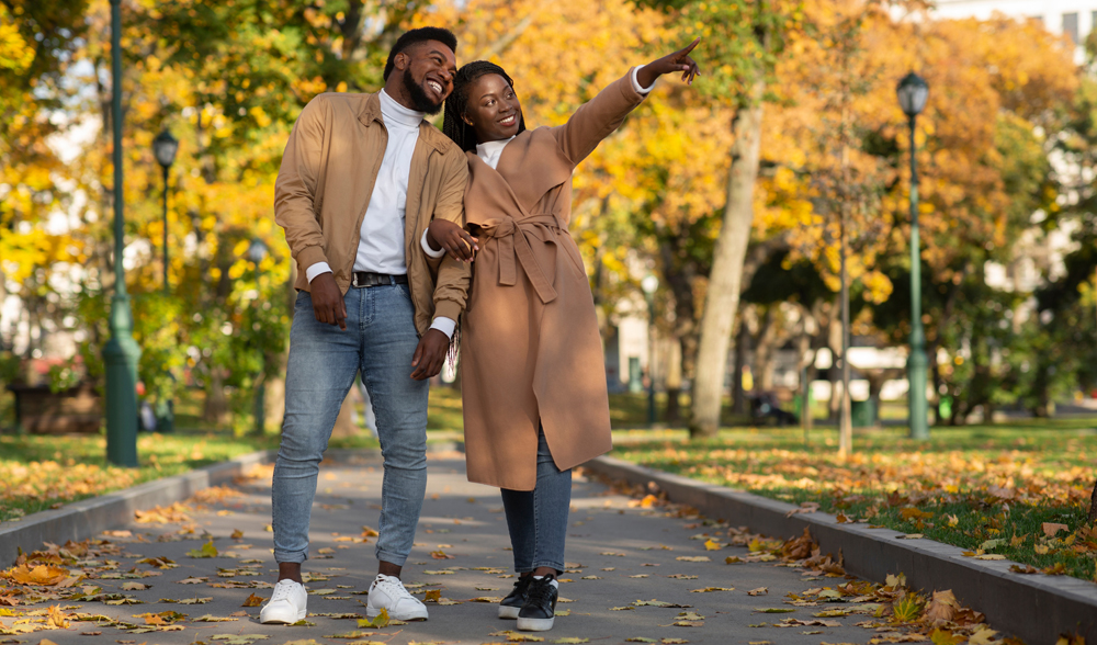 Black millennial couple walking on alley in autumn park, enjoying beauty of fall season, cheerful girlfriend pointing at something with finger