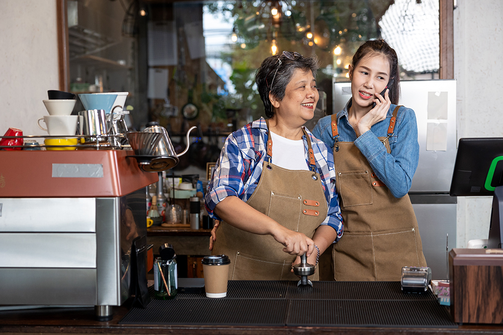 Asian senior and young woman working together at coffee bar. Elder woman smiling as younger talks on mobile phone. Generational teamwork and modern communication in family-run cafe business.